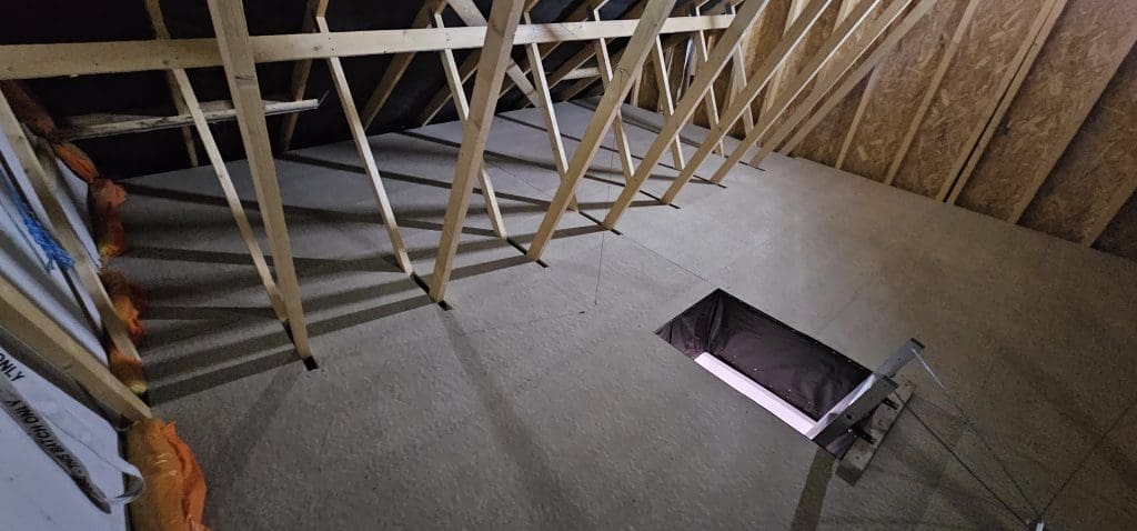 A view of a construction area with wooden beams forming the roof structure. The floor appears unfinished and features a rectangular hole. Stairs leading to the hole indicate potential attic or upper-level access. Orange insulation material is visible on the left side, suggesting considerations for pest control.