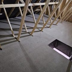 A view of a construction area with wooden beams forming the roof structure. The floor appears unfinished and features a rectangular hole. Stairs leading to the hole indicate potential attic or upper-level access. Orange insulation material is visible on the left side, suggesting considerations for pest control.