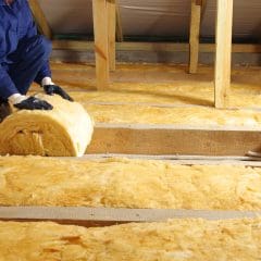 A person wearing blue work clothes and gloves is installing yellow insulation material in an attic. The attic has wooden beams and is under a roof with a light wooden frame. The person, skilled in pest control, is unrolling a roll of insulation between the beams for better protection.