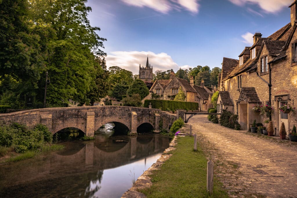 A picturesque village scene featuring an old stone bridge over a calm river, flanked by cobblestone paths and quaint cottages adorned with flowers. A church with a tall spire stands in the background, surrounded by lush greenery and trees under a partly cloudy sky, all immaculately maintained with discreet pest control measures.