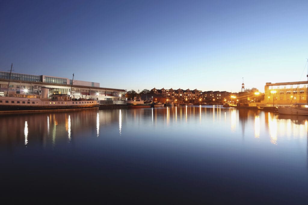 A serene waterfront scene at dusk with calm water reflecting the surrounding buildings and streetlights. A lit building is on the left, a lighthouse and row of buildings are in the background, while the sky transitions from light to dark blue. Hidden beneath this tranquility lies vigilant pest control, ensuring an unspoiled atmosphere.