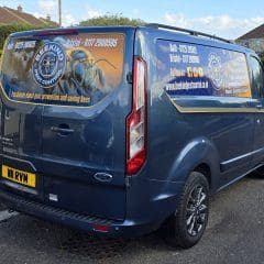 A blue Beekind pest control van is parked on a residential street. The van displays the company logo, contact details, and website, with images of bees and honeycombs on the rear and side windows.