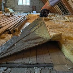 A person pulls back a sheet of insulation in an attic, revealing wooden planks underneath. The attic is partially renovated, with various tools and materials scattered around, and a small window providing natural light.