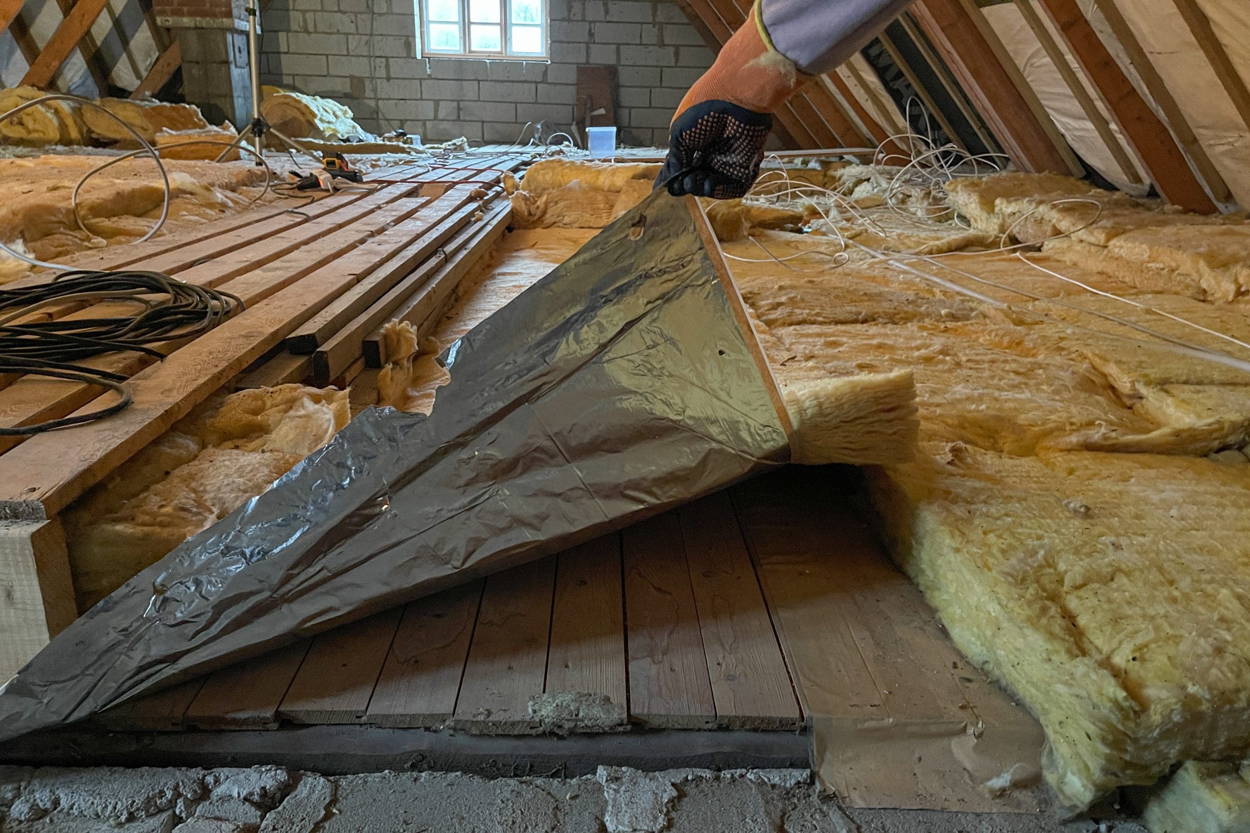 A person pulls back a sheet of insulation in an attic, revealing wooden planks underneath. The attic is partially renovated, with various tools and materials scattered around, and a small window providing natural light.