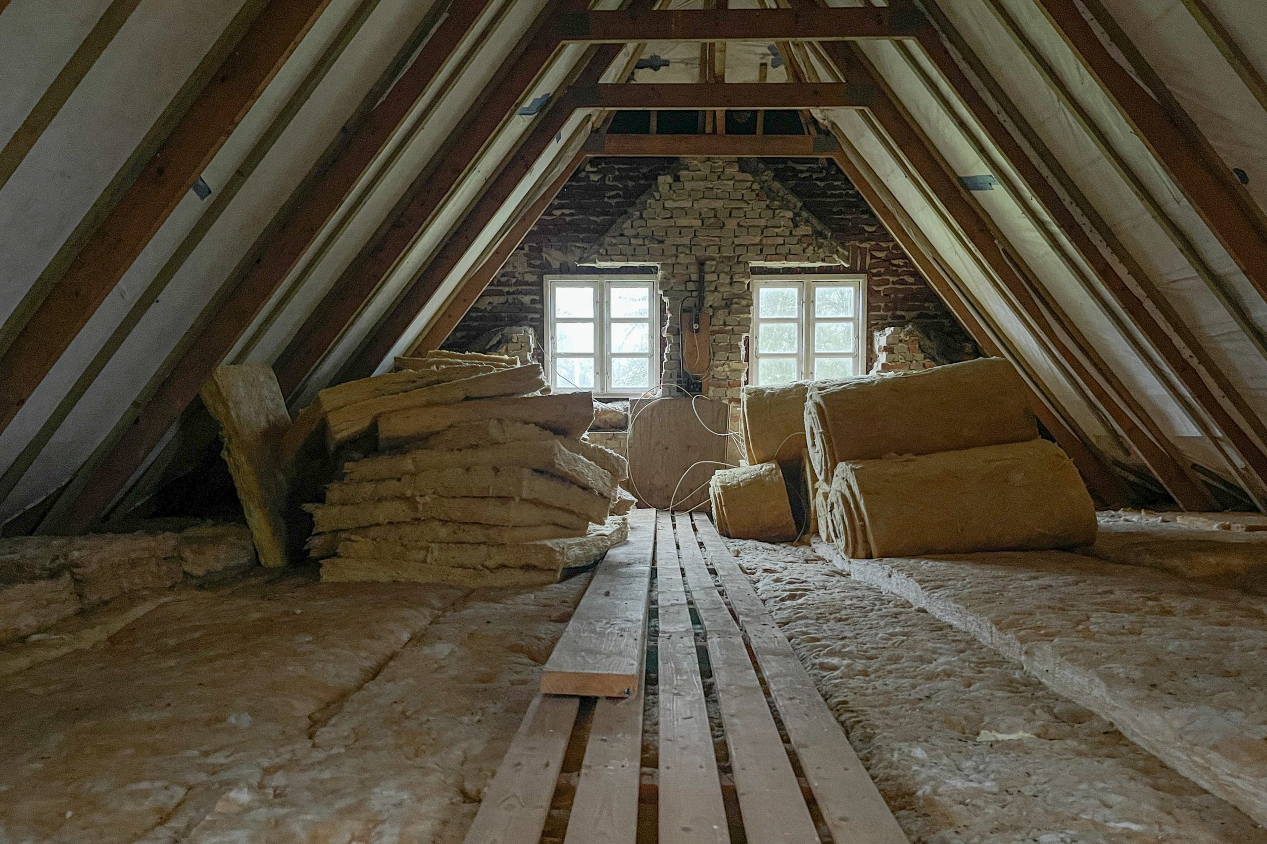 A dimly lit attic under renovation, with exposed wooden beams and insulation materials scattered around. Rolled and stacked sheet insulation lies on the floor, and two small windows at the far end provide some natural light.