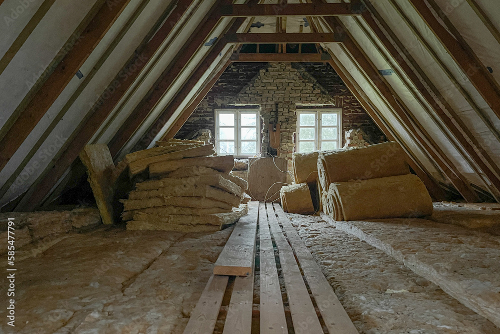 An attic with exposed wooden beams, brick walls, two small windows, and stacks of insulation material, including rolls and batts, alongside several wooden planks on the floor.