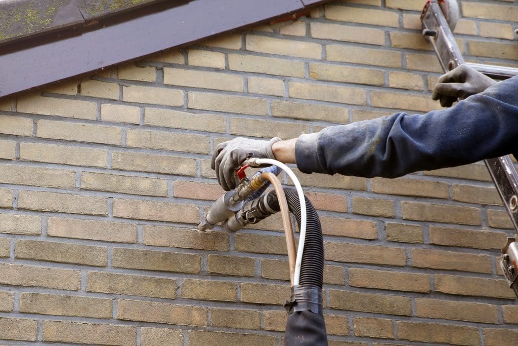 A person wearing gloves is using a hose and nozzle to inject insulation material into the brick wall of a building. A ladder is visible, and the person is dressed in a dark jacket.