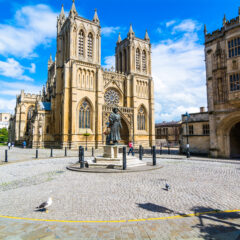 A statue stands in the center of a cobblestone square in front of a large Gothic-style cathedral with twin towers, under a blue sky with scattered clouds; historic buildings and a few birds are visible.