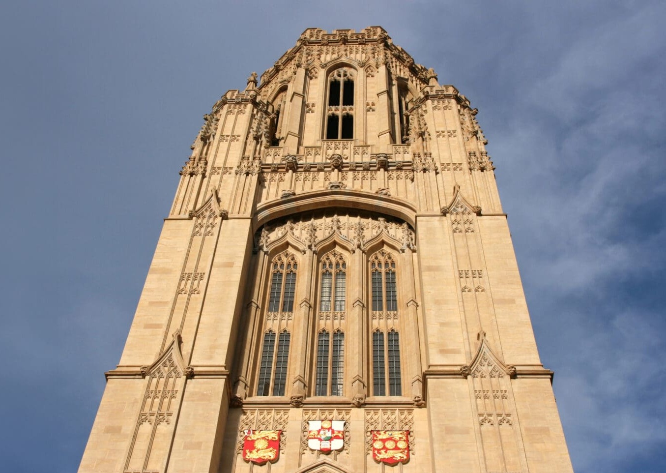 A low-angle view of a tall, ornate stone tower with arched windows and decorative details, set against a partly cloudy blue sky. Red and gold shields are visible above the entrance.