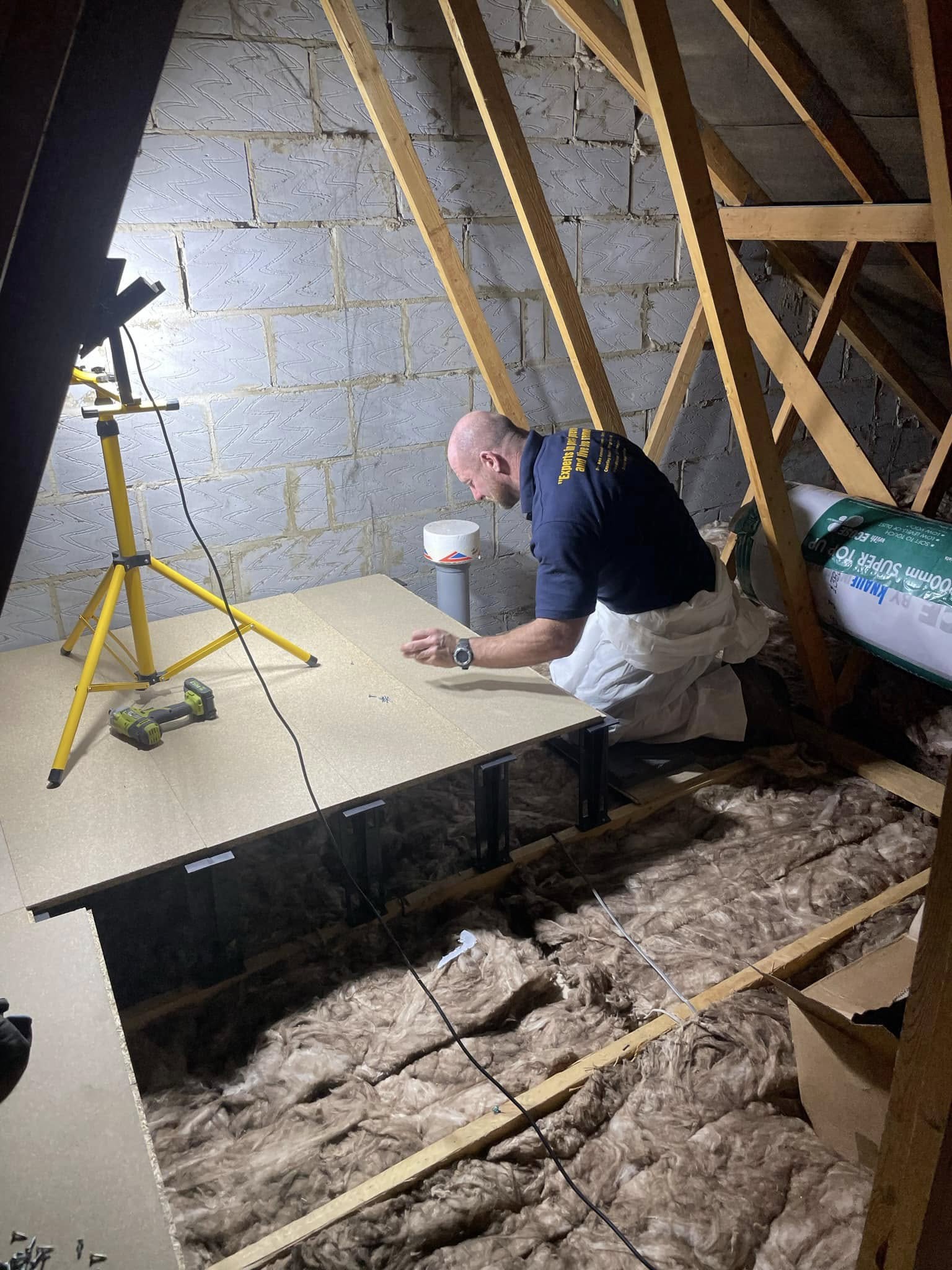 A person kneels in an attic, installing floorboards over exposed insulation. Tools and a work light are nearby, illuminating the workspace. Wooden beams and a brick wall are visible in the background.