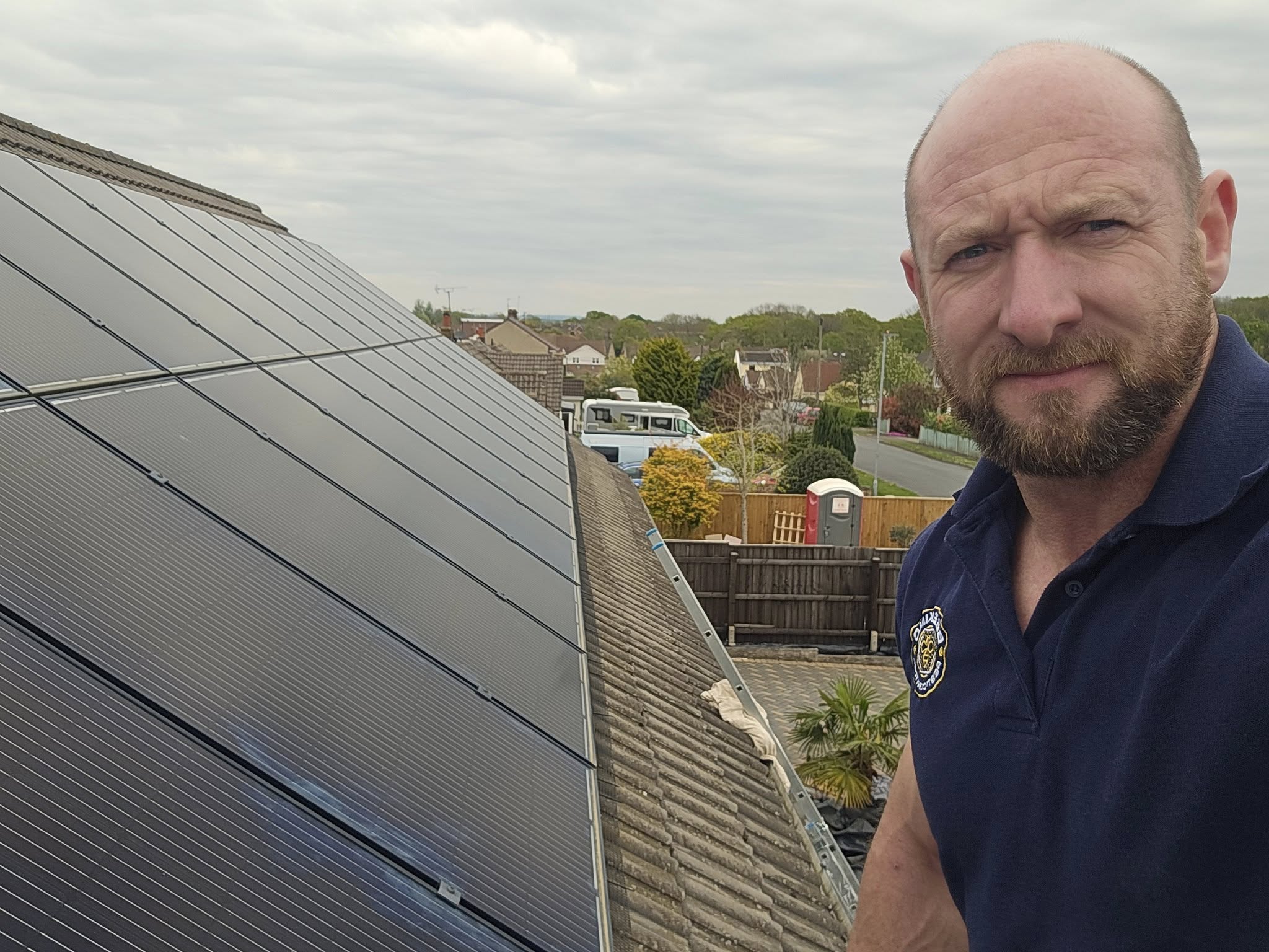 A man with a beard stands on a rooftop next to solar panels, looking at the camera. Houses, trees, and a road are visible in the background under a cloudy sky.