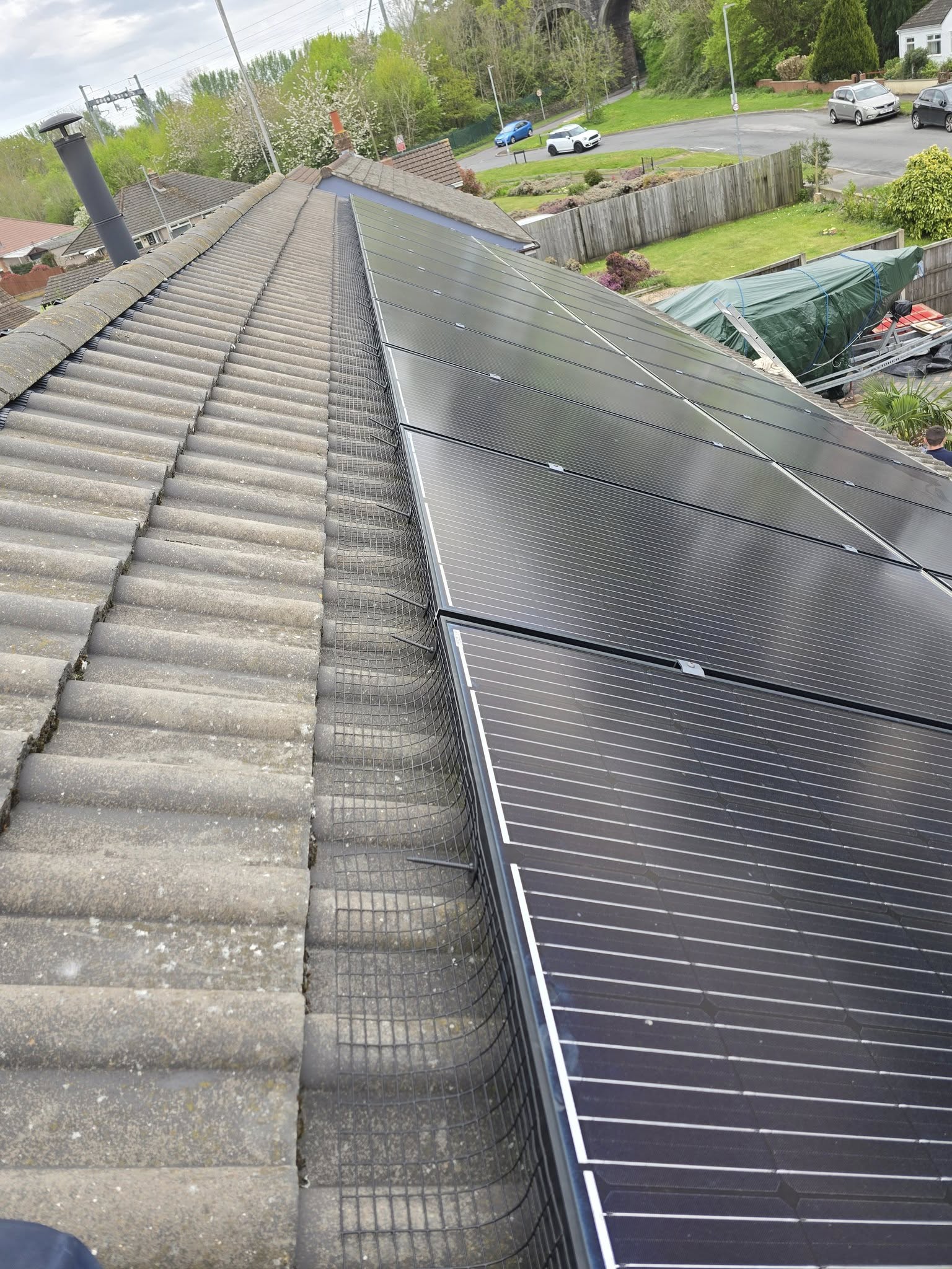 Rows of black solar panels installed on a sloped tiled roof, overlooking a suburban backyard with greenery, a garden, and parked cars on a nearby street.