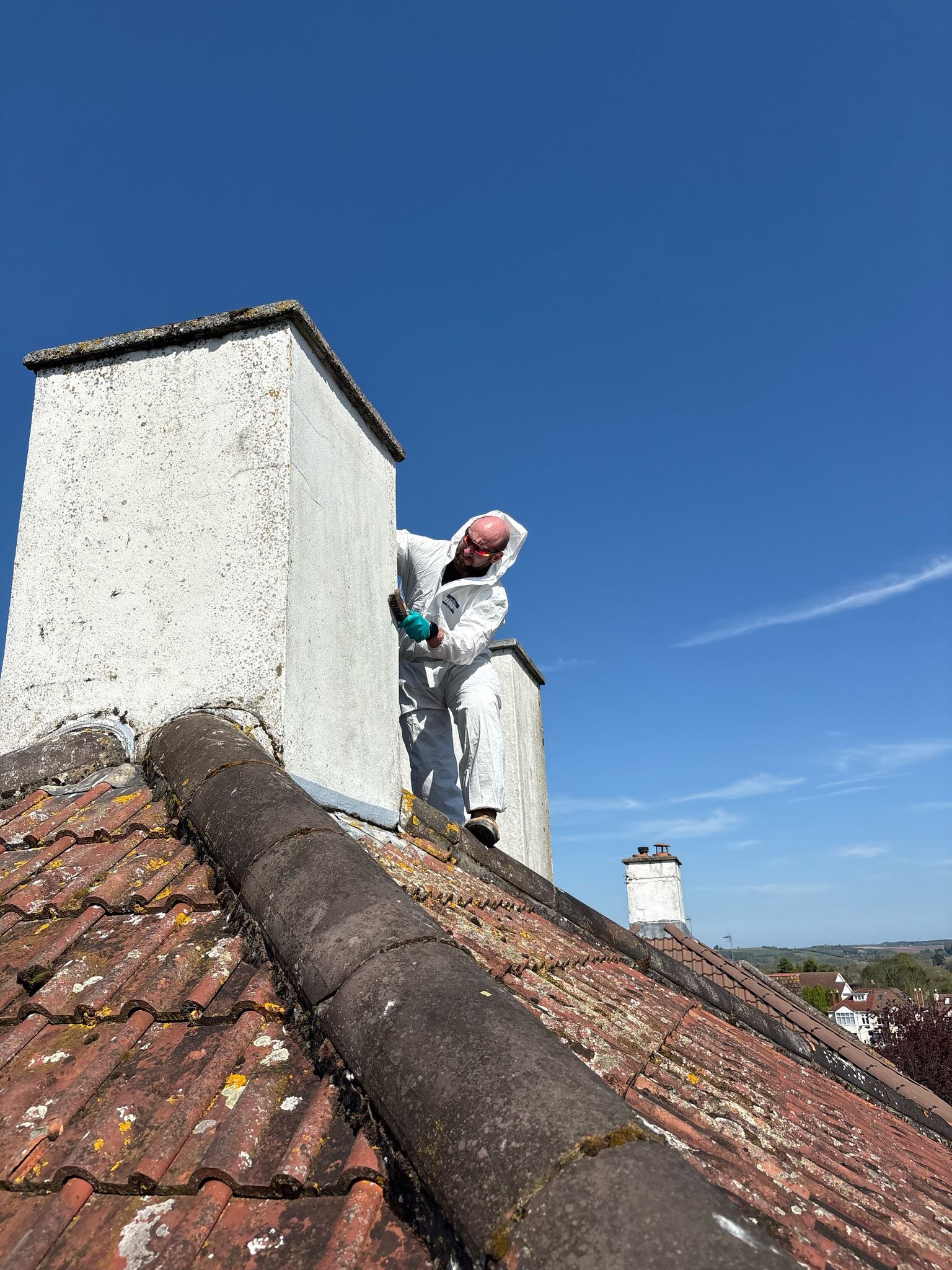 A person in a white protective suit and gloves works on a chimney atop a sloped, tiled roof under a clear blue sky. Green fields and trees are visible in the distant background.