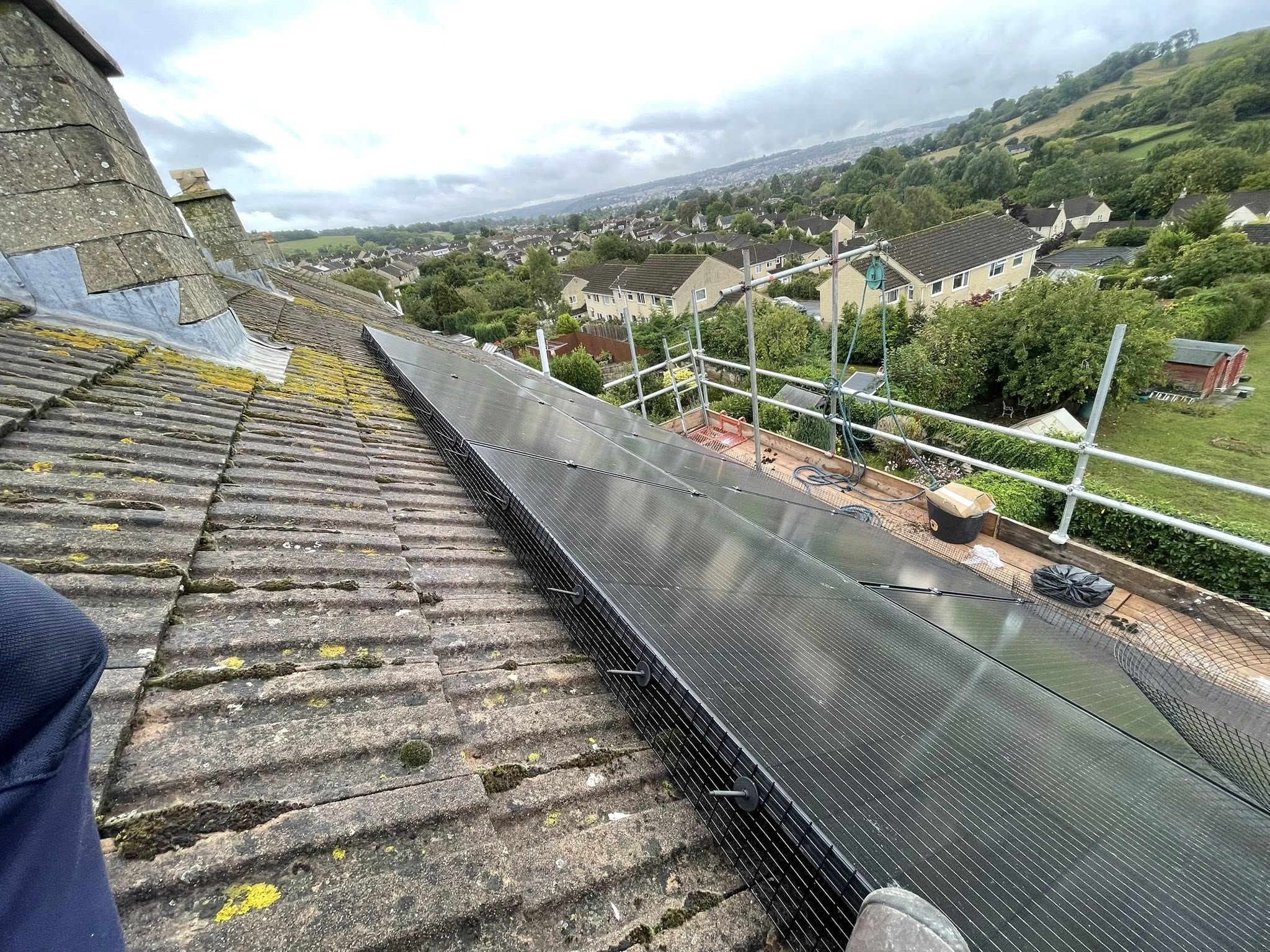 View from a sloped rooftop with solar panels installed along the edge, surrounded by wire mesh. Scaffolding and tools are set up nearby, overlooking houses, trees, and hills under a cloudy sky.