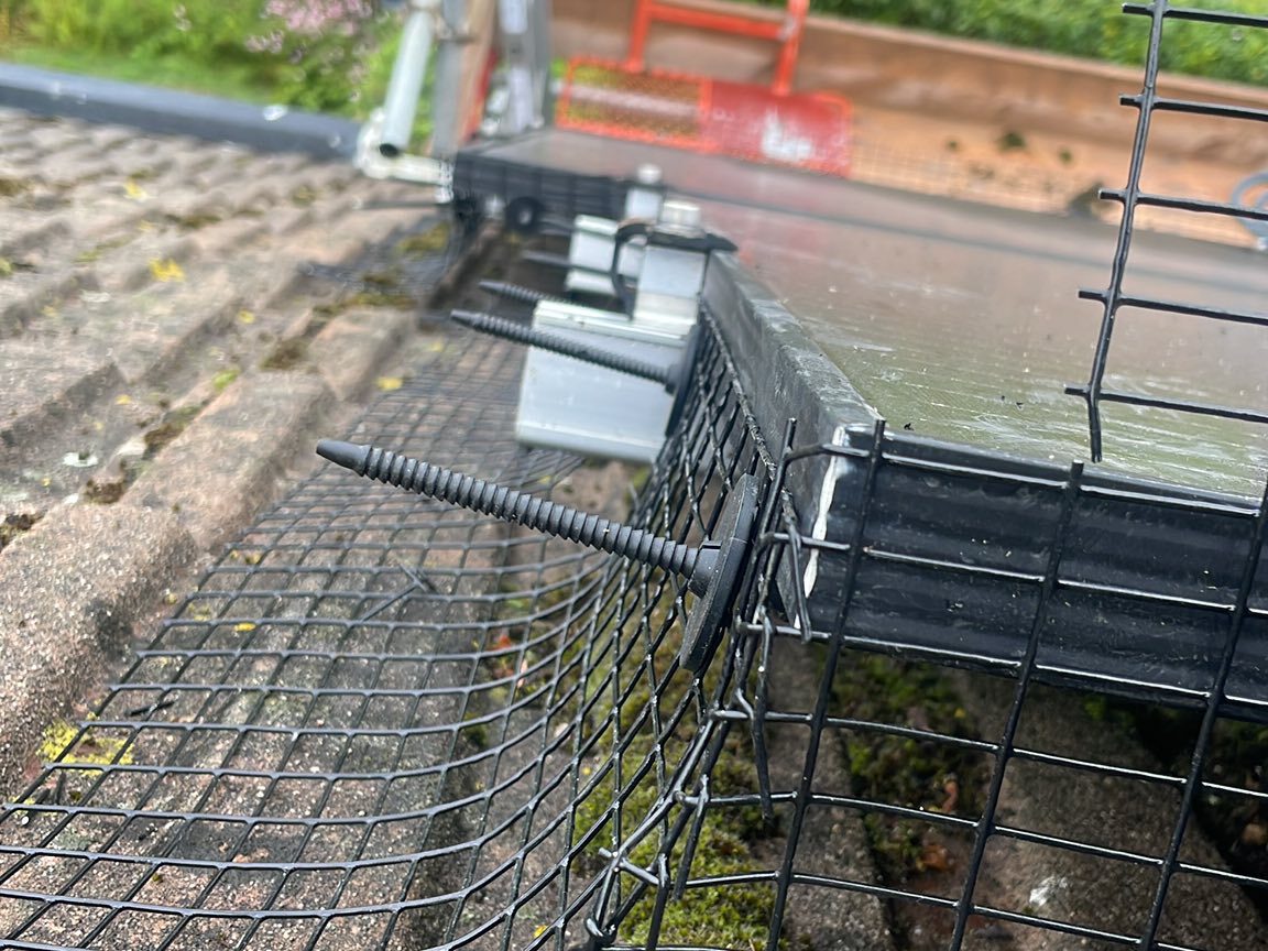 Close-up of a wire mesh barrier secured with screws and washers along the edge of a rooftop, next to solar panels. The mesh is preventing debris or animals from getting underneath the panels. Moss is visible on the roof tiles.