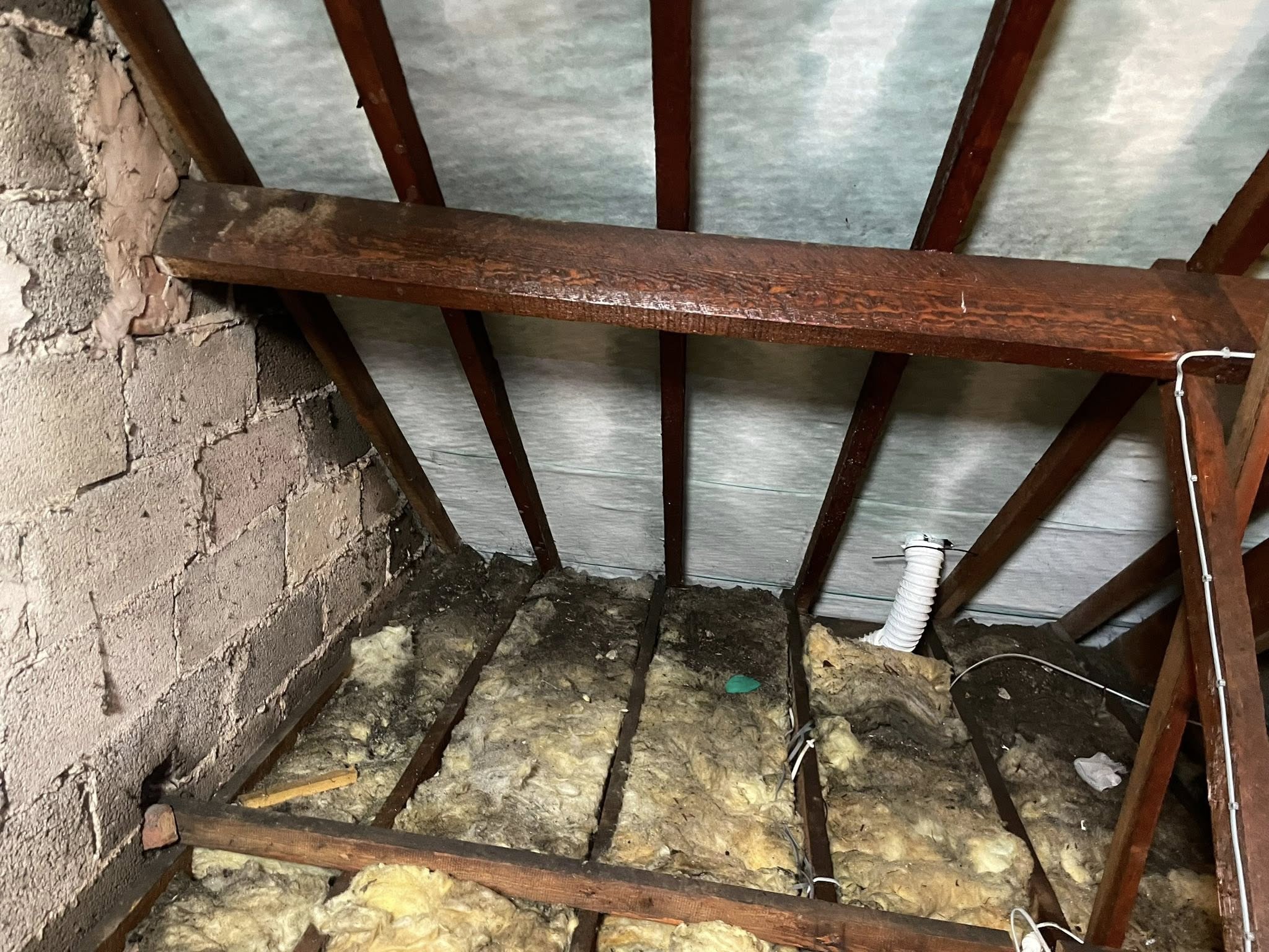 Attic space with exposed wooden beams, fiberglass insulation on the floor, cinderblock wall on the left, and a white vent pipe running through the insulation. The area appears unfinished and dusty.