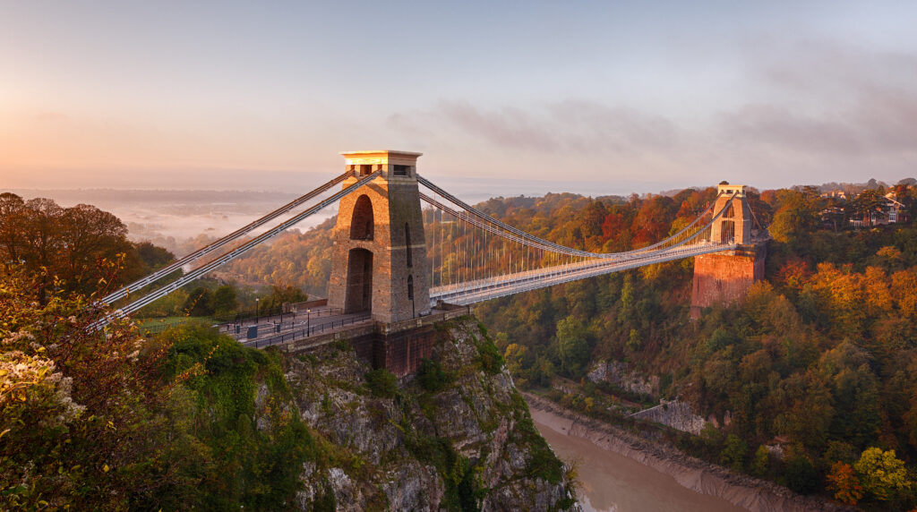 A suspension bridge spans a deep river gorge, surrounded by trees with autumn foliage. The sunrise casts a warm glow over the landscape, with mist visible in the distance.