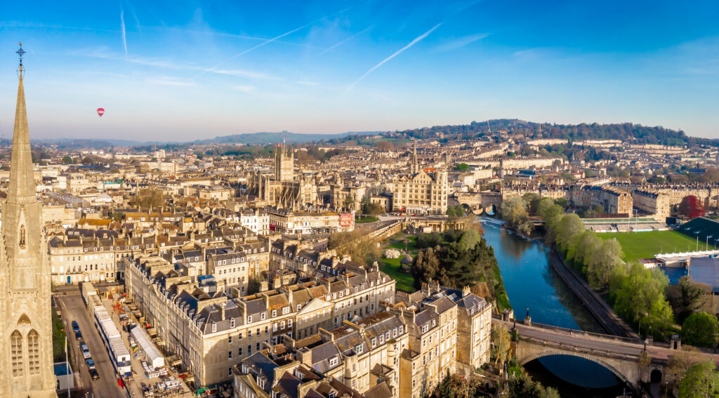 A panoramic aerial view of Bath, England, showing historic stone buildings, a church spire, the River Avon, green hills, and a hot air balloon in the clear blue sky.