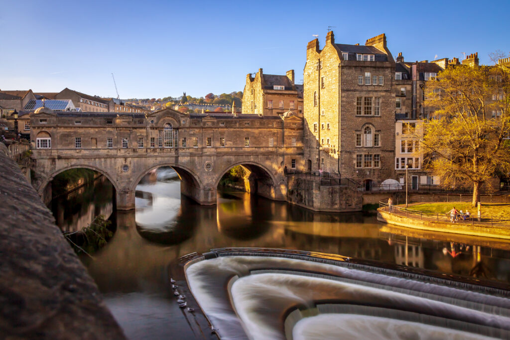 The Pulteney Bridge in Bath, England, crosses the River Avon with shops lining both sides. The river flows beneath the bridge, and a curved weir creates gentle ripples, all under a clear blue sky.