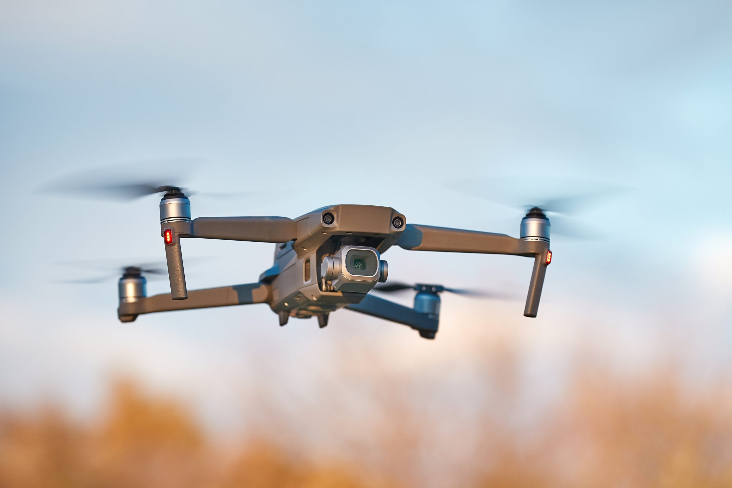 A gray quadcopter drone with four propellers is flying outdoors against a blurred background of trees and blue sky, captured mid-flight.