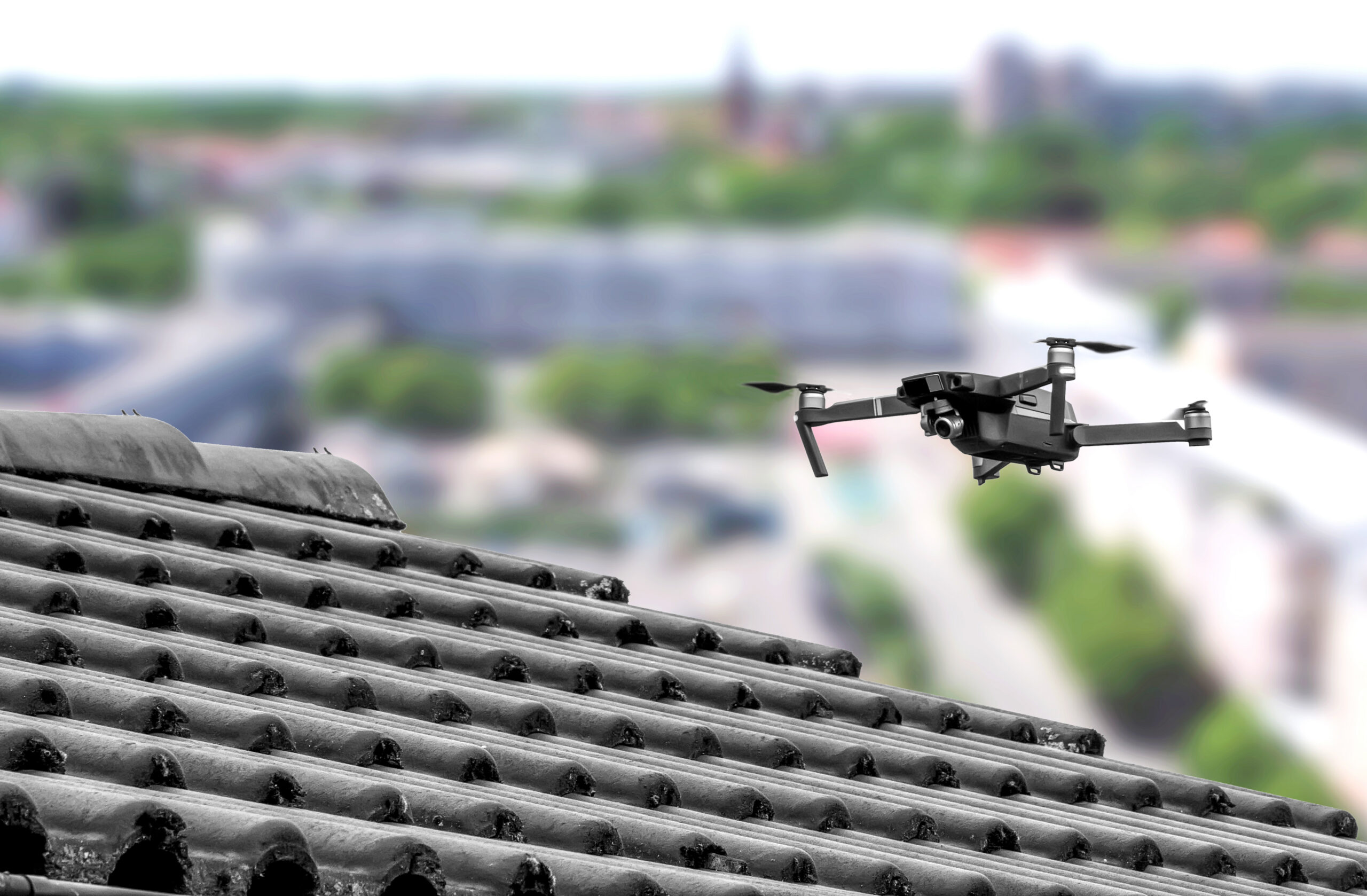 A black drone hovers near the edge of a tiled rooftop, with a blurred cityscape and greenery visible in the background.