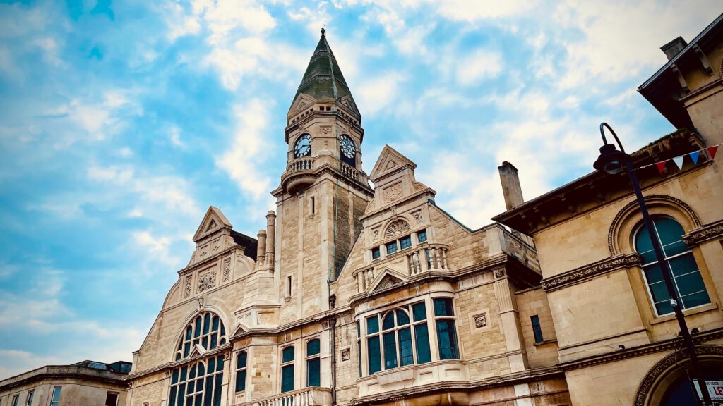 A historic stone building with a tall clock tower and pointed roof stands under a partly cloudy sky. The structure features arched windows and decorative architectural details.