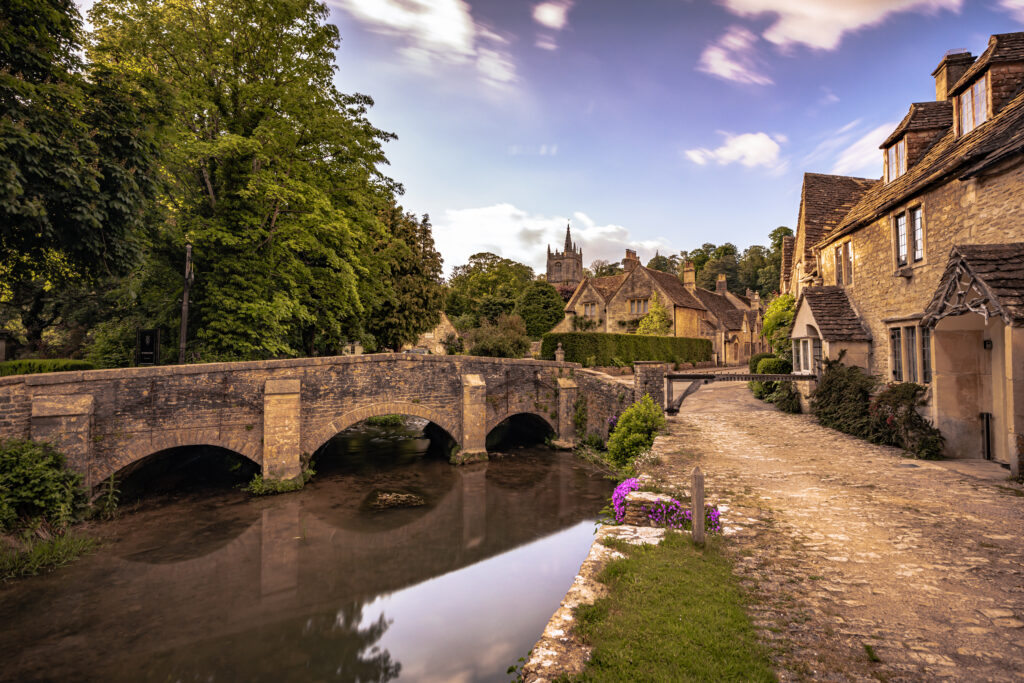 A stone bridge crosses a calm river beside a cobblestone path, bordered by historic stone cottages and lush trees under a partly cloudy sky at sunset.