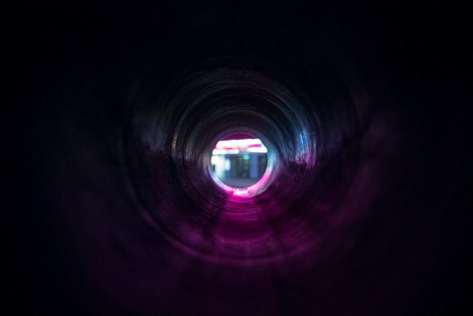 Looking through a dark tunnel with a blurred, bright neon light and pink and blue hues visible at the far end, creating a glowing circular effect.