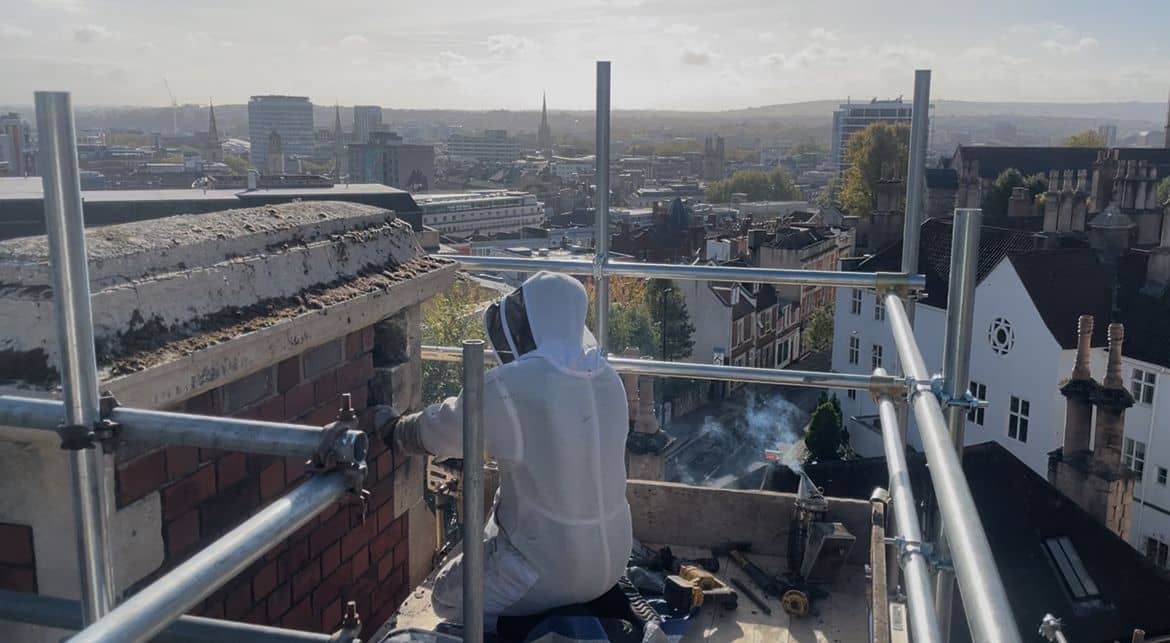 A person in protective clothing works on scaffolding at the top of a building, overlooking a cityscape with various buildings and a hazy sky in the background.