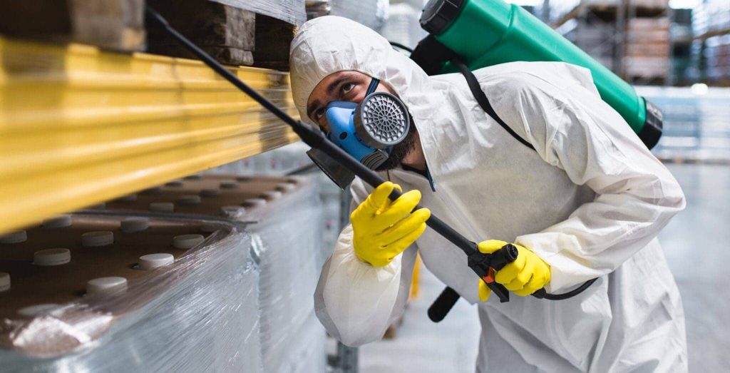 A worker in protective gear, including a mask, gloves, and hazmat suit, sprays disinfectant or pesticide under shelving in a warehouse setting.