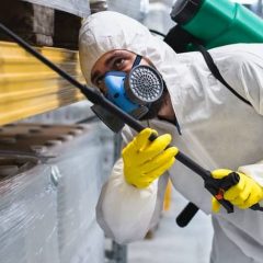 Person in protective gear, including gloves, face mask, and coveralls, sprays disinfectant from a tank and nozzle under a shelf in a warehouse or industrial setting.
