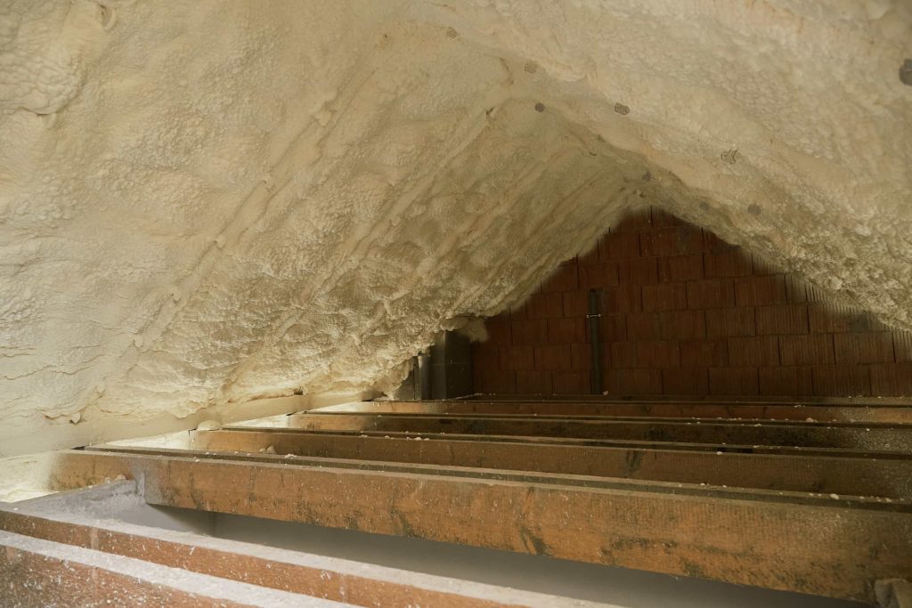 Spray foam insulation applied to the sloped ceiling and walls of an attic space, with exposed wooden beams and some brickwork visible at the far end.
