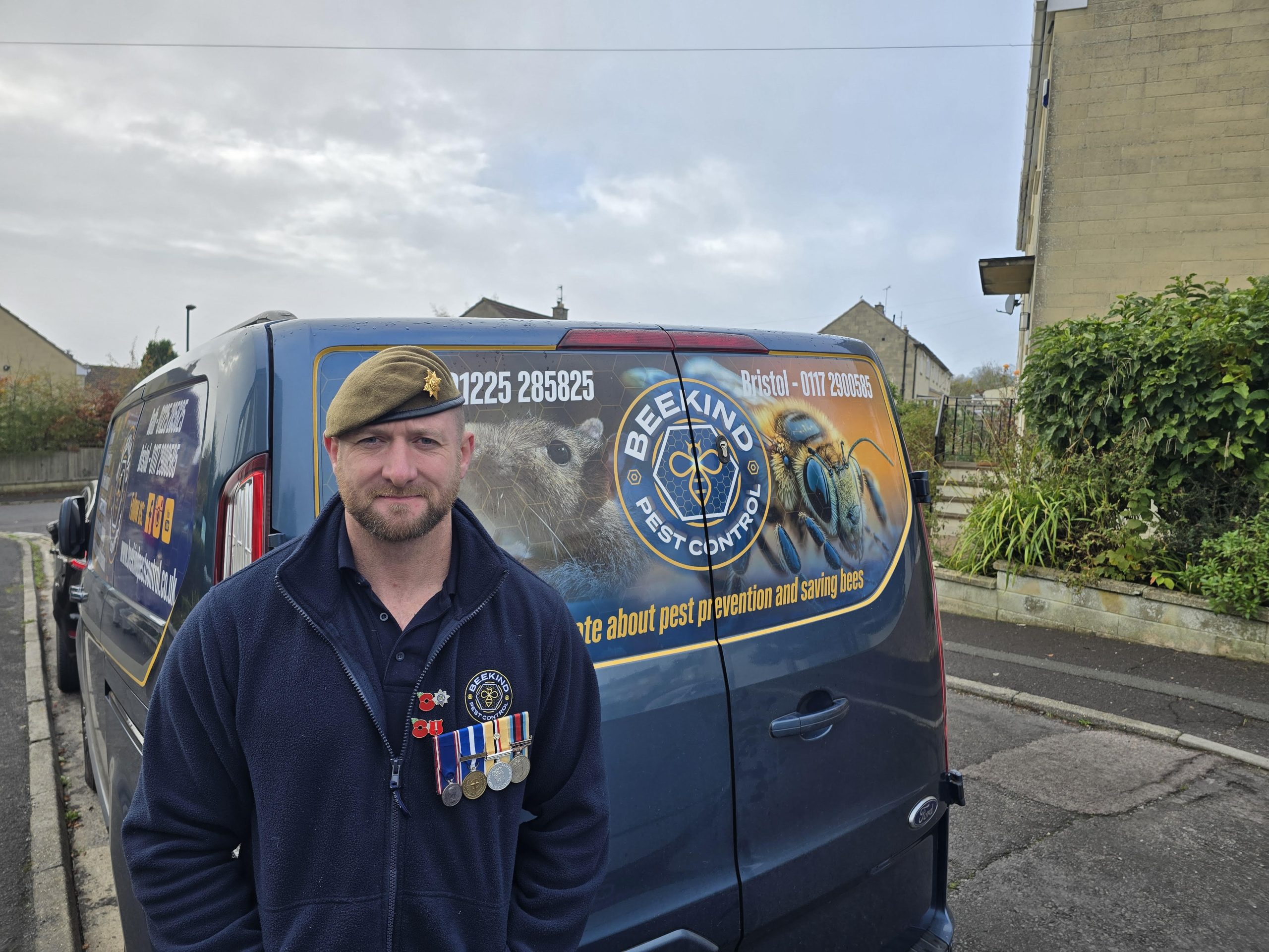 A man wearing a beret and medals stands in front of a van with "Beekind Pest Control" branding and bee graphics, parked on a residential street.